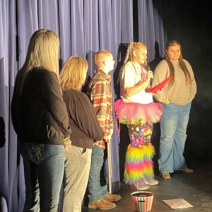 Five people standing center stage in front of a curtain. Fourth person from the left is holding a microphone and reading from a sheet of paper.