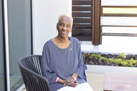 Portrait image of Maxine McMullen sitting outside in a wicker chair wearing a blue shirt.