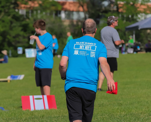 A man is playing cornhole in a park, wearing a blue T-shirt that says "Walking in Honor of My Wife" on the back. He is holding a bean bag, preparing to toss it towards a cornhole board. Other people and greenery can be seen in the background.