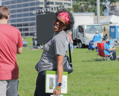 A person in a gray shirt, black pants, and red headband smiles while standing on a grassy field at an outdoor event. They hold a shopping bag with "Loving Life" on it and a binder. Other people, chairs, and equipment are in the background.