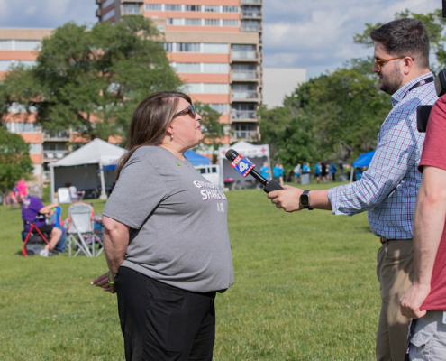 A woman in a gray shirt and black pants being interviewed by a man holding a microphone with the logo "FOX 4". They are standing on a grassy field with a tall building in the background, and people seated on lawn chairs in the distance.