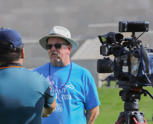 A man wearing a light grey hat and blue Legacy Walk 2024 t-shirt being interviewed