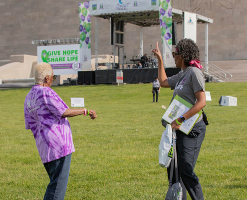 Two people engage in conversation on a grassy field in front of a stage with a "Give Hope Share Life" banner. The background features a building with engravings and steps leading to the stage.