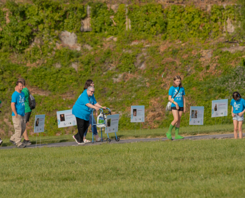 A group of people walk along a path with signs raised on stakes to their right. Most are wearing blue-green T-shirts and walking shoes. The signs display photographs and text. The area is grassy with a hillside covered in greenery in the background.