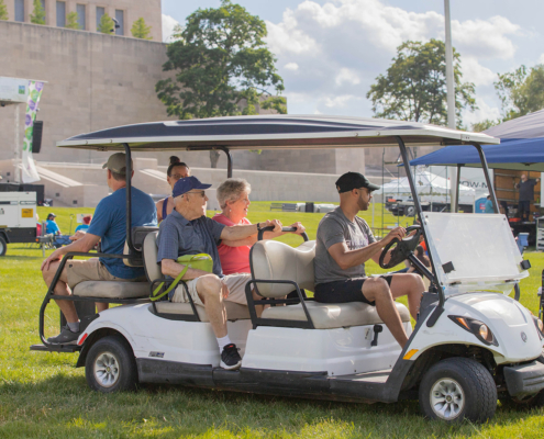 A man drives a six-seater golf cart with four elderly passengers through a grassy park on a sunny day. There are outdoor structures and event setups in the background.