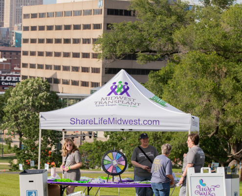 A group of people stand under a white tent with "Midwest Transplant Network" written on it. The tent has a spinning wheel on a table and informational materials. It's set up on a grassy area with city buildings in the background.