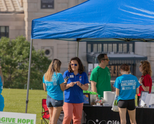 A group of people standing under a blue canopy tent at an outdoor event. The tent has a table with informational materials, and a sign nearby reads "Give Hope Share Life." The backdrop includes a large building.