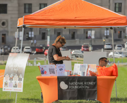 An orange tent with a table is set up on a grassy area. The table has a black National Kidney Foundation banner. A woman is writing while a man sits nearby, wearing sunglasses.