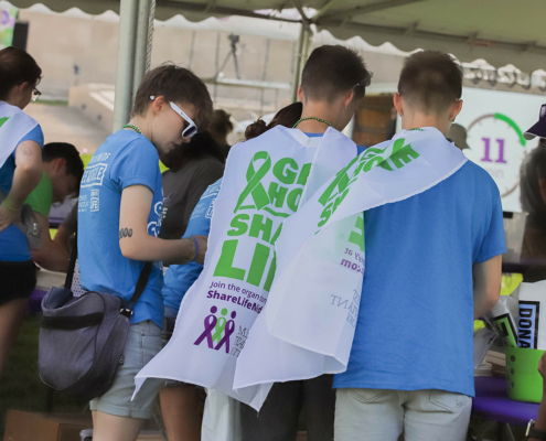 Three participants wearing capes are seen from behind under a tent at an outdoor event.