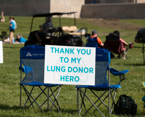 A blue folding chair on grass holds a sign that reads "Thank You To My Lung Donor Hero" in blue text. People are gathered in the background, with some sitting in folding chairs and others standing.