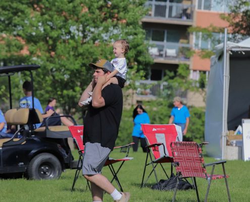 A man in a black T-shirt and gray shorts carries a child on his shoulders while walking on a grassy area. The child is smiling and wearing a white shirt. In the background, there are a few people in blue shirts and some chairs near a building with large windows.