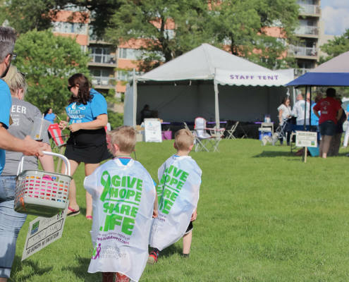 Two kids wearing capes on a grass field with tents in the background