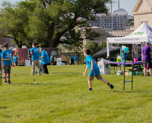 People at an outdoor event on a grassy field. Some are playing ladder toss, while others stand near a tent with "ShareLifeMidwest" banners. In the background are trees, a few buildings, and other participants engaging in various activities.