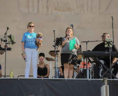 A band and people on an outdoor stage. Two people are holding microphones.