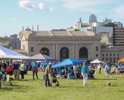 People at an outdoor event with tents and a city in the background on a sunny day.