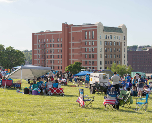 People at an outdoor event with tents and a building in the background on a sunny day.