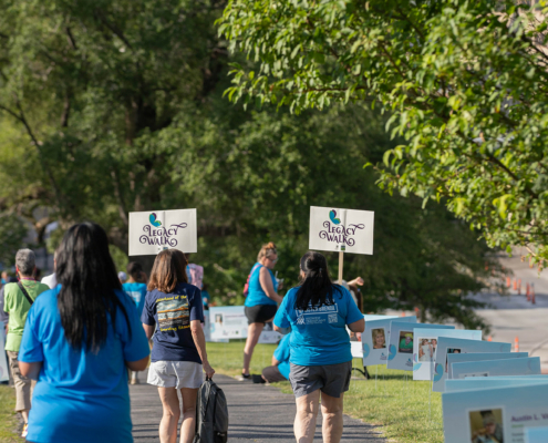 A group of people participates in an outdoor walkathon, viewed from the back. Several carry signs with the text "Legacy Walk." They are walking on a paved path with trees and green grass around them. The weather appears sunny and pleasant.