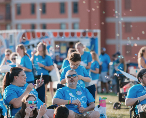 People sitting on chairs on a grass field with a food truck in the background. Some are blowing bubbles.