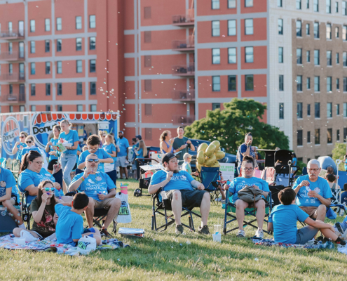 People sitting on chairs on a grass field with a food truck in the background. Some are blowing bubbles.