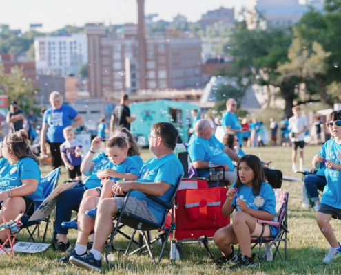 People sitting on chairs on a grass field with city buildings in the background. Some are blowing bubbles.