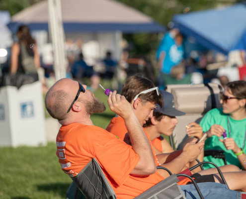 A man with a beard blows bubbles while sitting in a chair at an outdoor event. Several other people, some seated and some standing, are in the background.