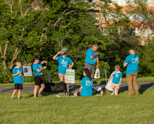 A group of six people, including children and adults, wearing matching blue shirts gather in a grassy area near signs. Some are standing, while others are sitting on the ground. Trees and a building in the background suggest an outdoor event or activity.