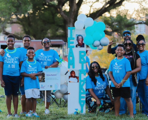 A group of people wearing blue Legacy Walk 2024 shirts stand together outdoors, smiling. They are gathered around a large sign that reads TEAM RICKEY.