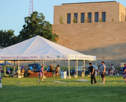 A group of people enjoying outdoor activities under a large white tent with a "Welcome to the Legacy Walk" banner.